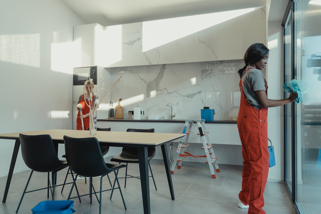 services-04 Two women in work clothes cleaning a kitchen with glass doors.